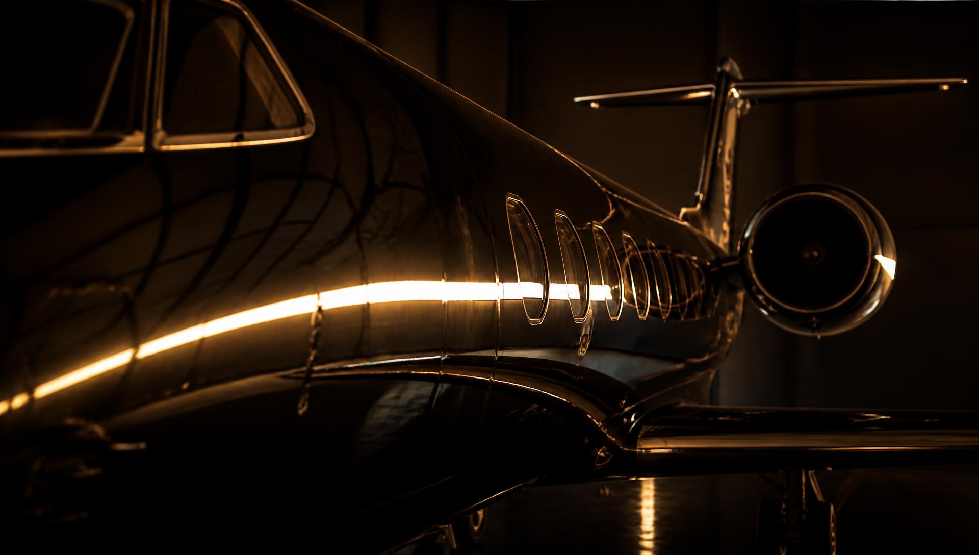Glossy private jet fuselage catching a band of warm light in a dark hangar